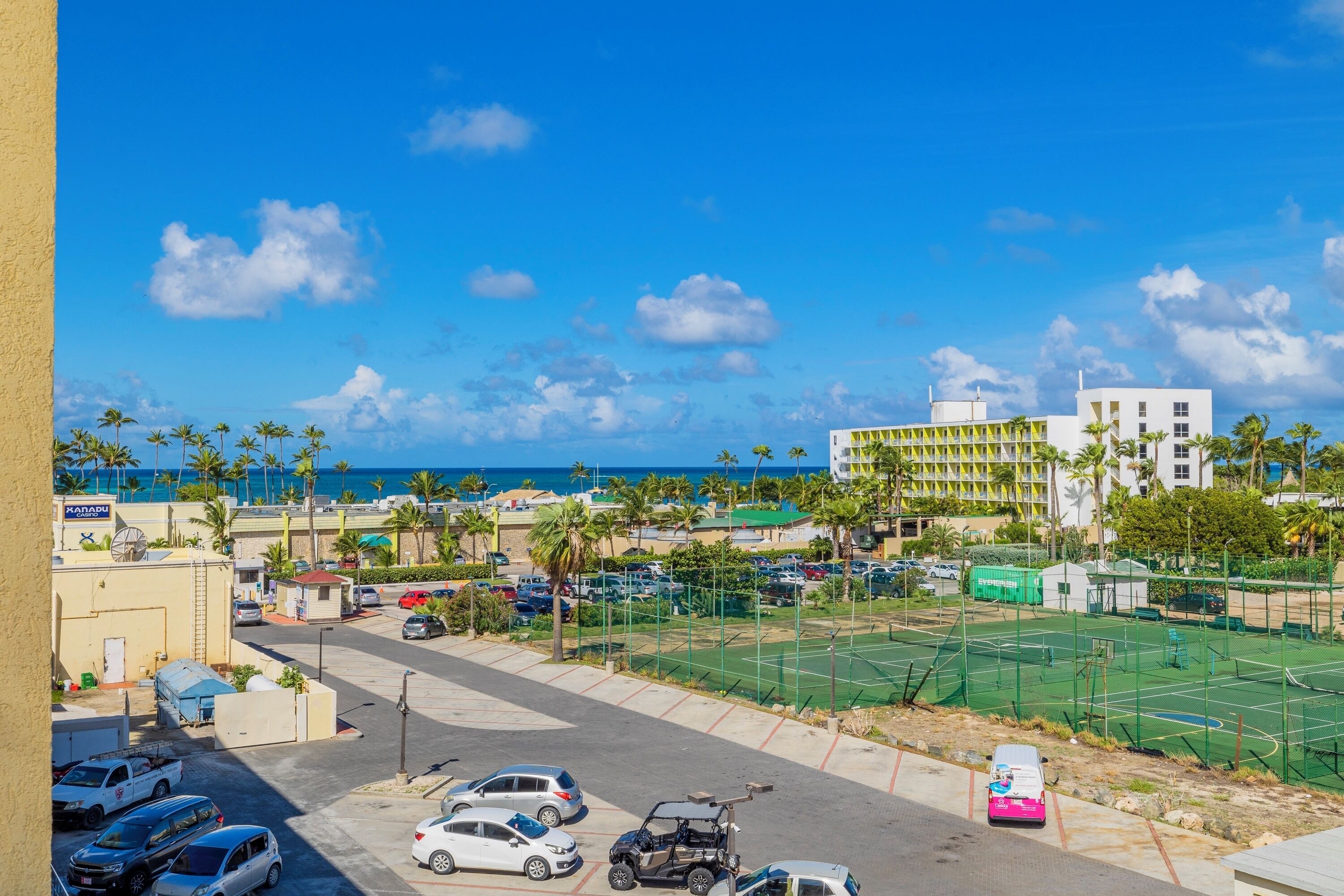A parking lot with several cars and a golf cart, a tennis court, and buildings with palm trees and a view of the ocean.