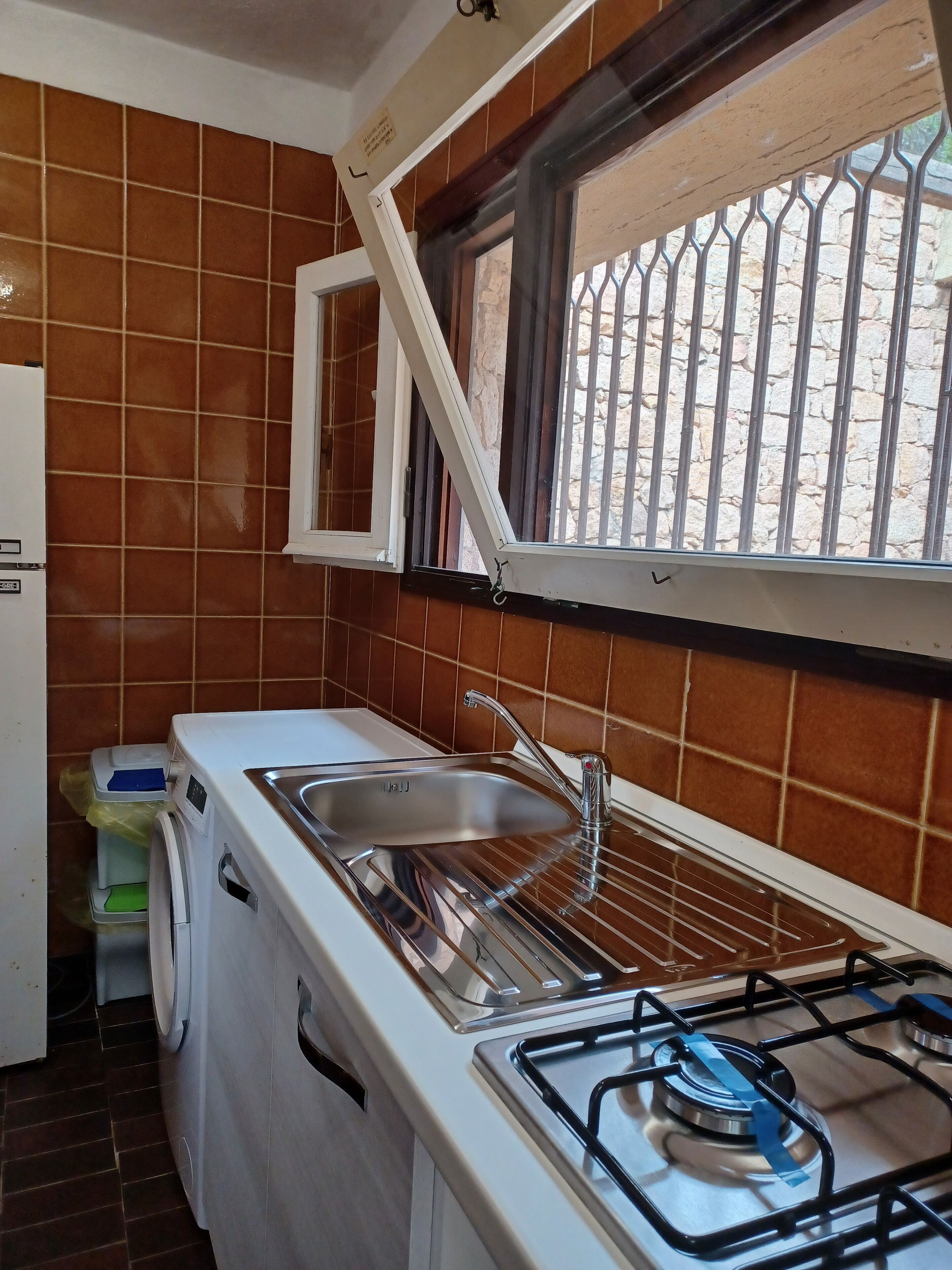 A compact kitchen with a sink, a gas stove, a washing machine, and a window with a metal grill.