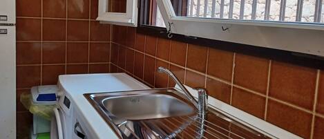 A compact kitchen with a sink, a gas stove, a washing machine, and a window with a metal grill.
