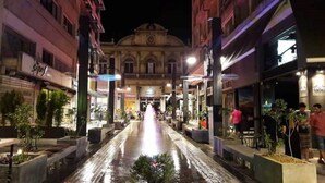 Vista nocturna de una calle con una fuente en el centro, flanqueada por edificios con comercios a ambos lados.