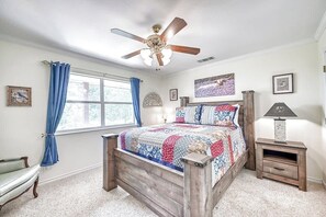 A bedroom with a wooden bed, a ceiling fan, a window with blue curtains, and a nightstand with a lamp. - Hill Country Oasis-Deck-BBQ Grill-10 Min to Town! (Fredericksburg)