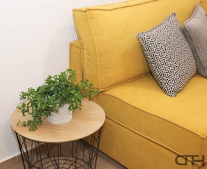 Un sofá color mostaza con cojines estampados, una mesita redonda con una planta en maceta y una base de metal negro.