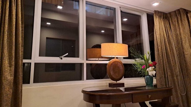 Ein Zimmer mit einem großen Fenster, einem Tisch mit einer Lampe und einer Vase mit Blumen.