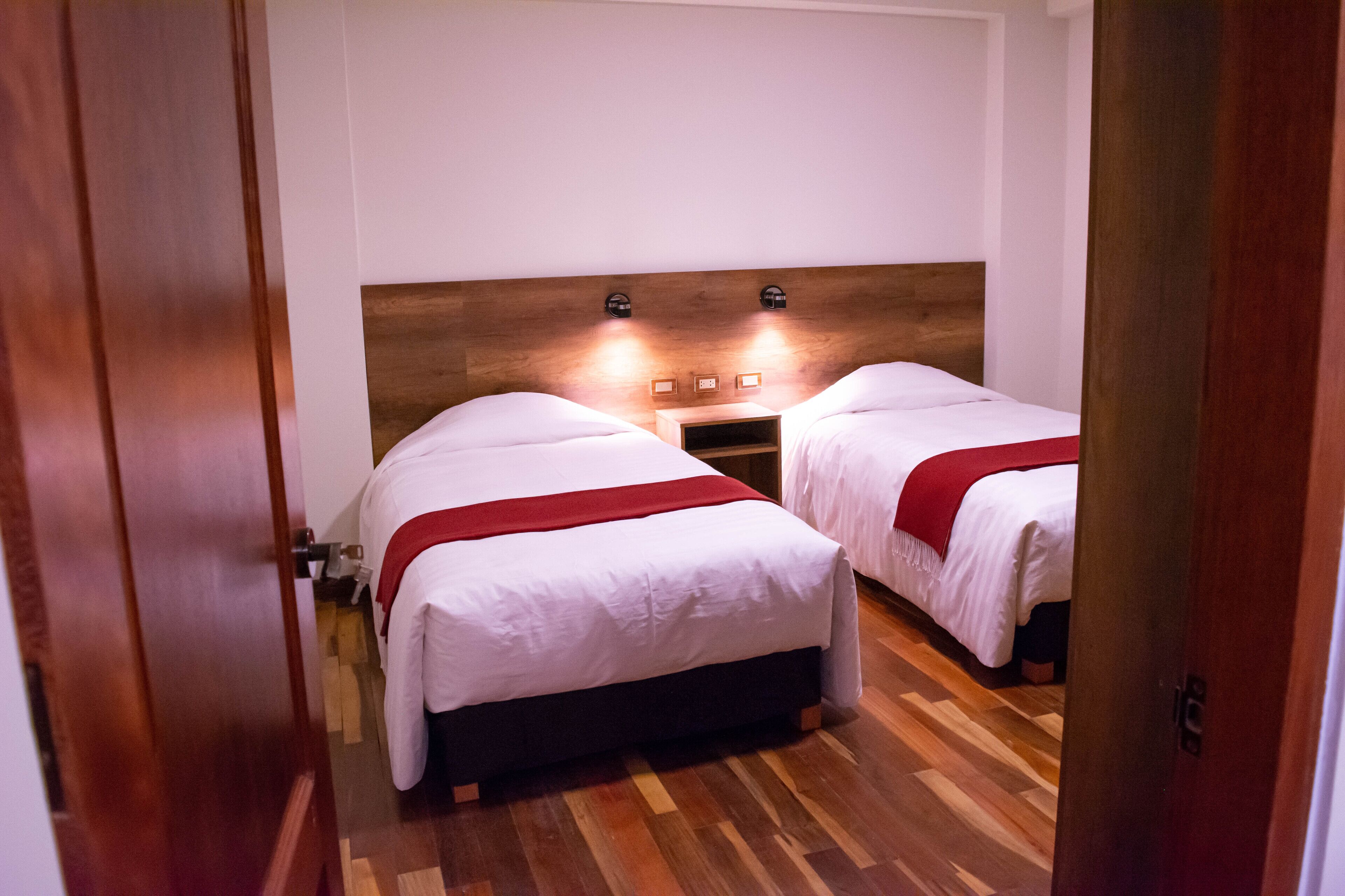A hotel room with two beds, wooden headboard, bedside lamps, and a wooden floor.