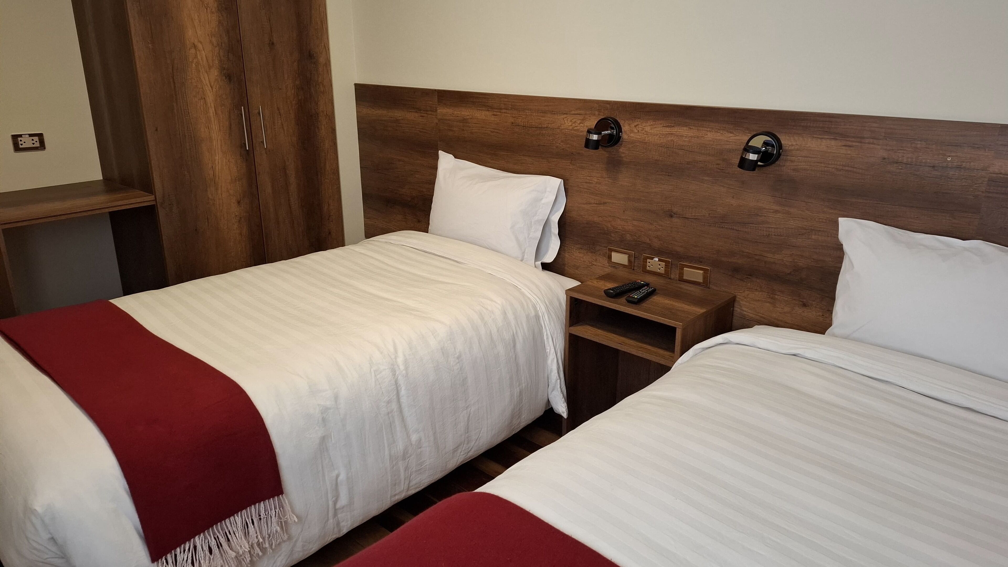 A hotel room with two beds, wooden headboards, a nightstand with a lamp, and a wardrobe.