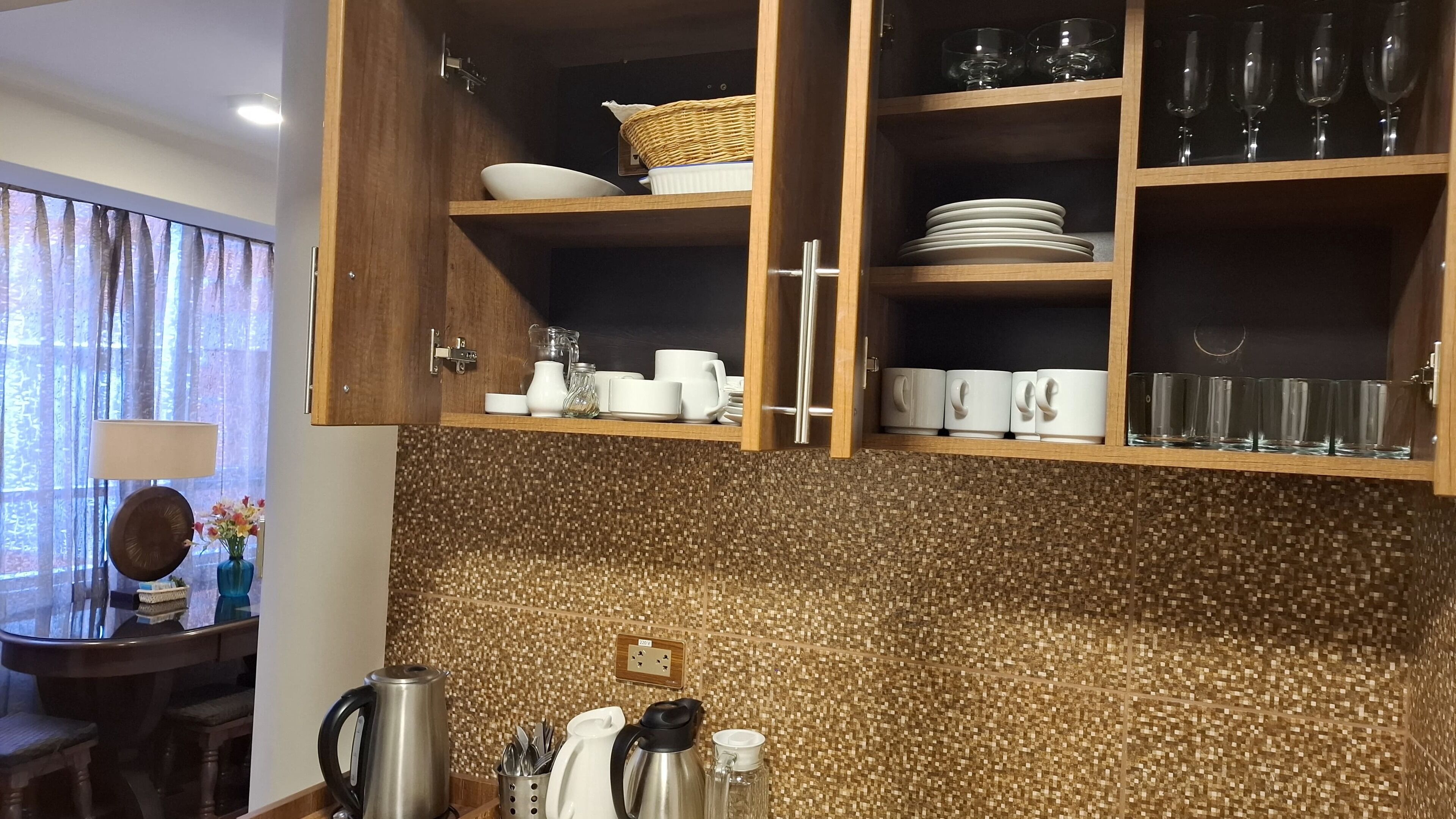 Uma cozinha com armários de madeira, painel de azulejos e diversos utensílios, incluindo chaleira, xícaras e pratos.