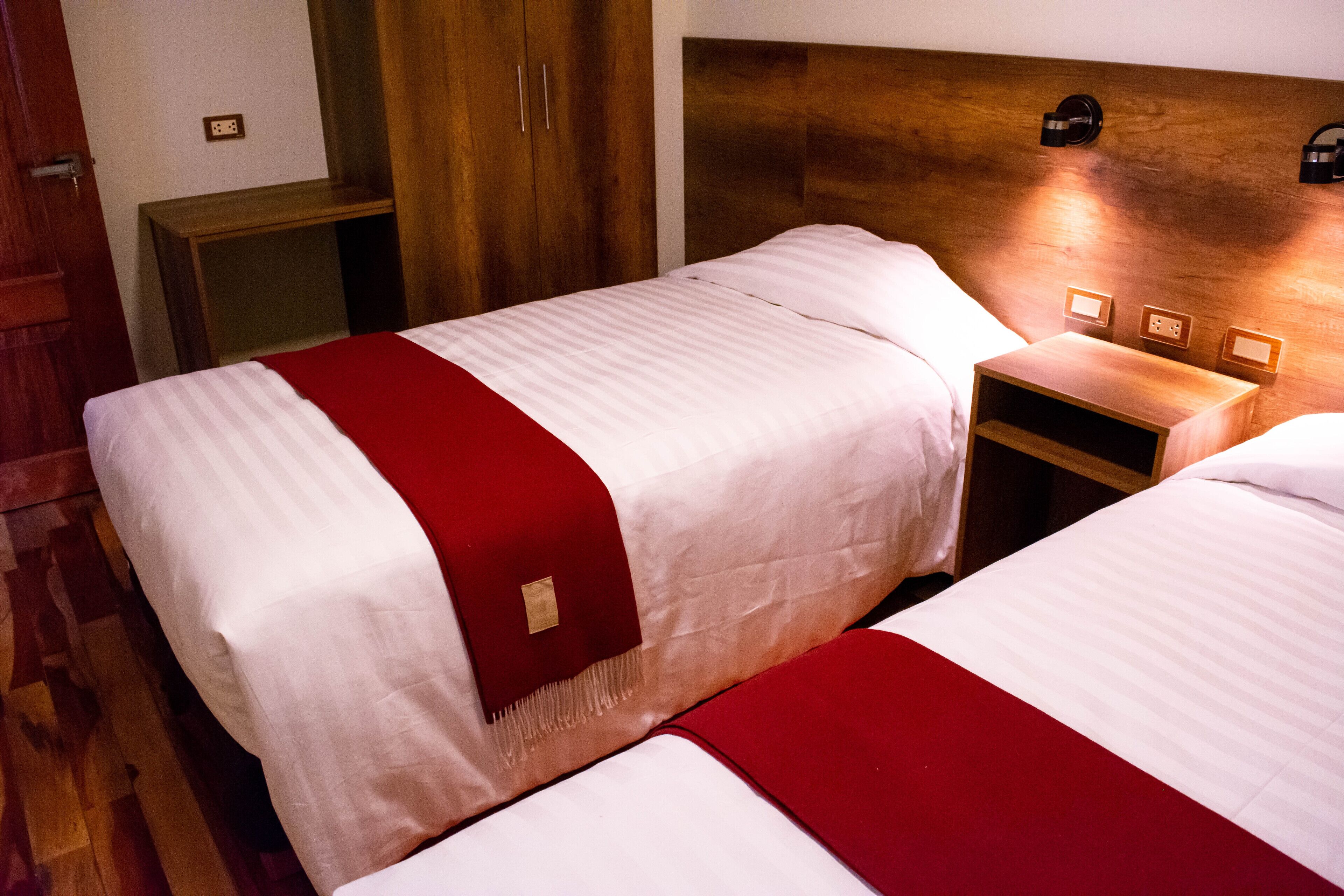 A hotel room with two beds, wooden headboard, bedside tables, and a red accent on the beds.