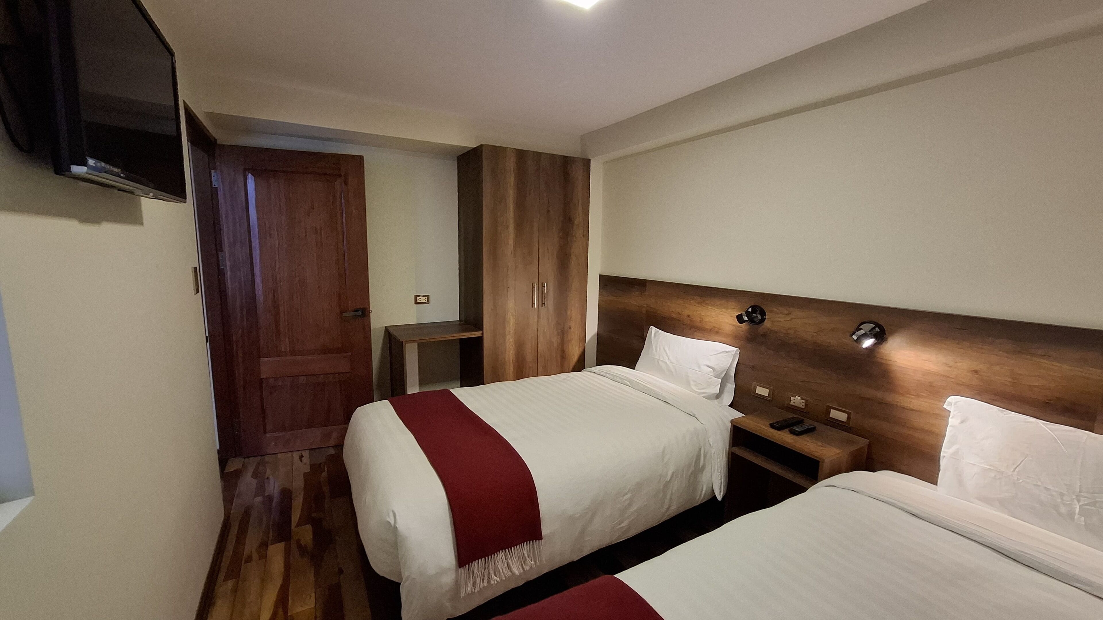 A hotel room with two beds, wooden wardrobe, bedside tables, and a TV mounted on the wall.