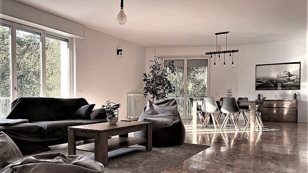 A modern living room with a sofa, coffee table, and bean bag chairs. There is a dining area with a table and chairs. A large window provides natural light, and a painting is visible on the wall. - Spacious and bright apartment surrounded by greenery 10 minutes from Varese (Valganna)