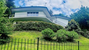A modern two-story house with a curved balcony, surrounded by lush greenery and a green fence. - Spacious and bright apartment surrounded by greenery 10 minutes from Varese (Valganna)