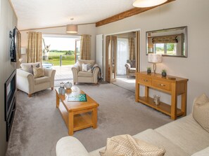 A spacious living room with a sofa, armchairs, a coffee table, and a wooden side table. There is a television mounted on the wall, and a large window provides a view of the outdoors. - Stewarts Resort Lodge 29 - a lodge that sleeps 6 guests in 2 bedrooms (St Andrews)