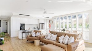 Een moderne woonkamer met een beige bank, een salontafel en een eethoek met een tafel en stoelen. Achtergrond is een keuken met witte kasten en roestvrijstalen apparatuur.