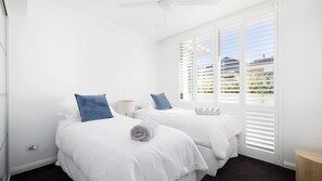 Een hotelkamer met twee bedden, wit beddengoed en uitzicht door grote ramen met jaloezieën.