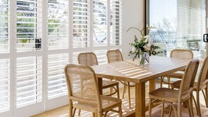 Een eetruimte met een houten tafel en rieten stoelen, grote ramen met jaloezieën en een vaas met bloemen op de tafel.