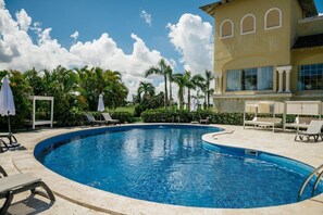 Ein Swimmingpool mit Liegestühlen und Sonnenschirmen, umgeben von Palmen und einem Gebäude mit Bogenfenstern.