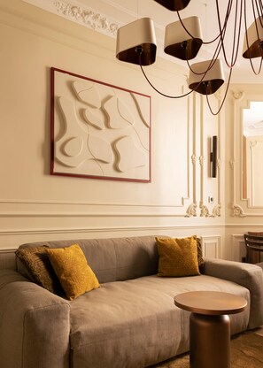 A modern living room with a beige sofa, two mustard-colored pillows, a round coffee table, and a wall-mounted abstract art piece. - Luxury Suite - Champs Elysees - Wagram (Paris)