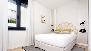 Une chambre à coucher avec un lit, une fenêtre avec des rideaux, un luminaire fixé au mur et un tableau encadré.