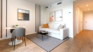 Ein modernes Wohnzimmer mit einem weißen Sofa, einem grauen Sessel, einem runden Couchtisch und einem kleinen Beistelltisch. Eine weiße Wand ziert den Raum, darüber befindet sich ein gerahmtes Kunstwerk, und links an der Wand hängt ein Spiegel.