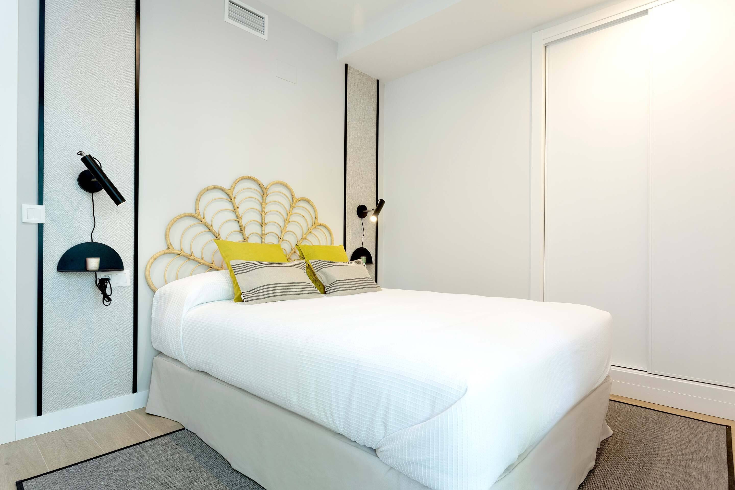 Una cama bien hecha con edredón blanco y almohadones decorativos, una lámpara de pared y una puerta blanca sencilla en una habitación moderna.