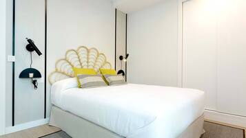 Una cama bien hecha con edredón blanco y almohadones decorativos, una lámpara de pared y una puerta blanca sencilla en una habitación moderna.