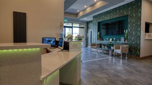 A reception area with a counter, two monitors, a seating area with chairs, a large TV on the wall, and a patterned wall.