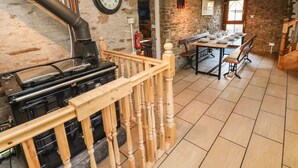 A rustic kitchen with a wood-burning stove, stone walls, and a dining area with a long table and benches. - Nant Moel Isaf Farm (Swansea)