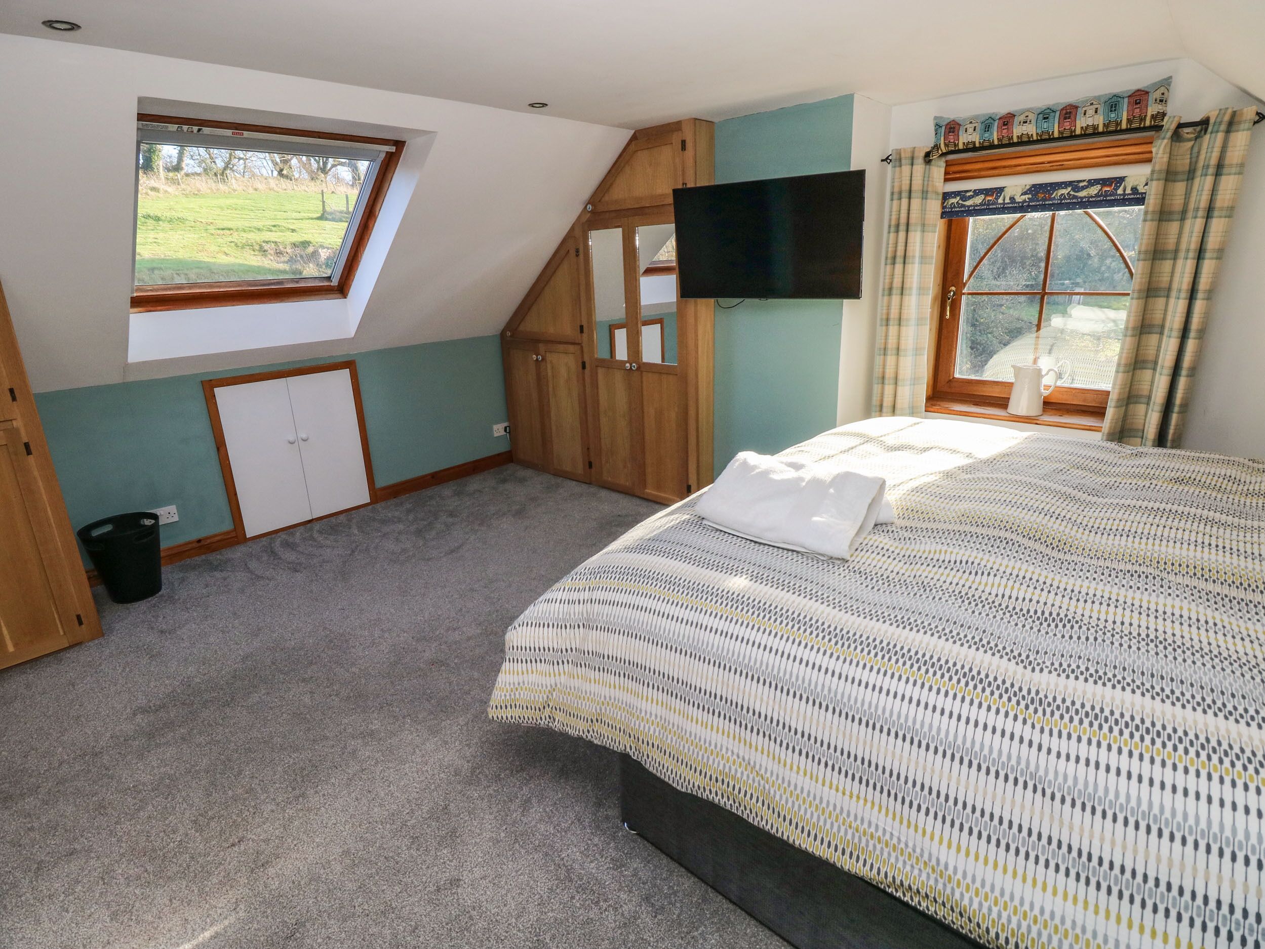 A bedroom with a sloped ceiling, a large window, a flat-screen TV, a bed with a patterned bedspread, and a small closet.