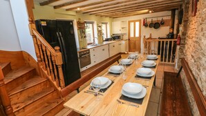 A dining area with a long wooden table set for a meal, a kitchen in the background, and a wooden staircase leading to another level. - Nant Moel Isaf Farm (Swansea)