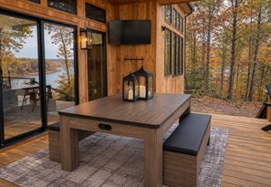 Eine Holzterrasse mit Tisch und Bank, einem an der Wand befestigten Fernseher und Blick auf Herbstbäume.