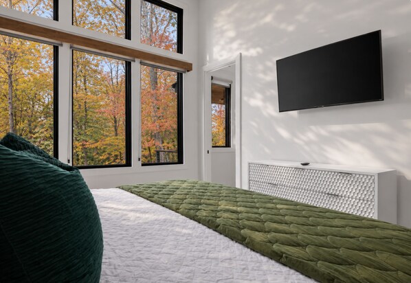 A modern bedroom with a large window, a bed with green and white bedding, and a flat-screen TV mounted on the wall.