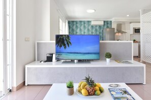 Un soggiorno moderno con una televisione a schermo piatto su una consolle bianca, un tavolo da pranzo con una ciotola di frutta e una rivista.