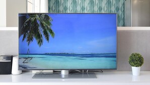 Litteänäyttöinen televisio valkoisella jalustalla, pieni ruukkukasvi ja lautanen täynnä hedelmiä valkoisella pöydällä.