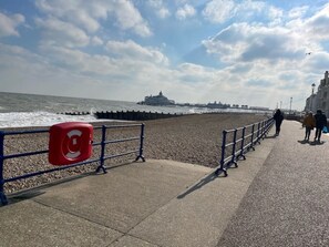 A seaside promenade with a pebbled beach, a pier in the distance, and a red safety sign on the railing.