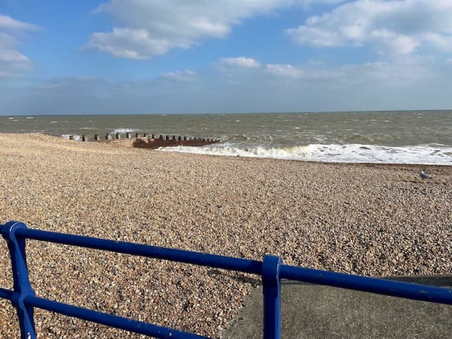 Une plage de galets avec une rambarde bleue au premier plan, des vagues qui se brisent sur le rivage et un ciel nuageux.