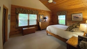 A bedroom with wooden walls, a ceiling fan, a bed, a dresser, and two windows with blinds. - The Cabin at Denali Stud (Paris)