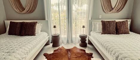 Une chambre avec deux lits, un grand tapis marron et une fenêtre ornée de rideaux légers.