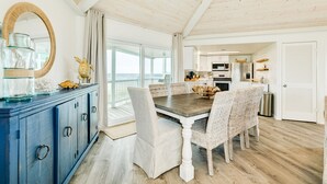 Ett rymligt matområde med ett träbord och flätade stolar, ett blått skåp med glasvaror och utsikt över havet genom stora fönster.
