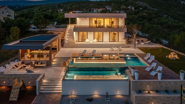 Une maison moderne à deux étages dotée d’une grande piscine extérieure, d’un patio couvert et offrant une vue sur les collines environnantes.