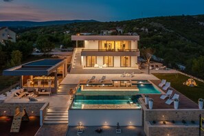 Una casa moderna de dos pisos con una amplia piscina exterior, un patio cubierto y vistas a las colinas circundantes.