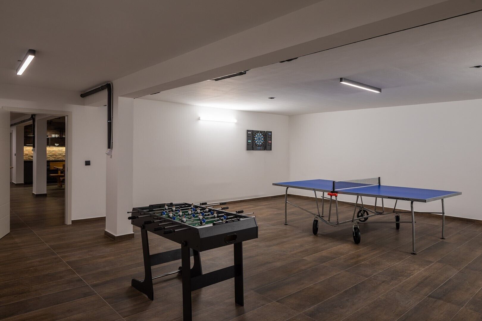 A room with a foosball table, ping pong table, and dartboard.