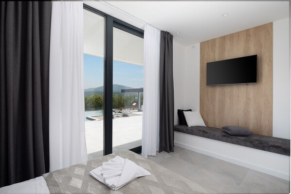 Une chambre d’hôtel moderne dotée d’une grande porte-fenêtre coulissante, d’un téléviseur à écran plat fixé sur un mur en bois, d’un canapé avec des coussins et d’une serviette blanche soigneusement pliée posée sur le sol.