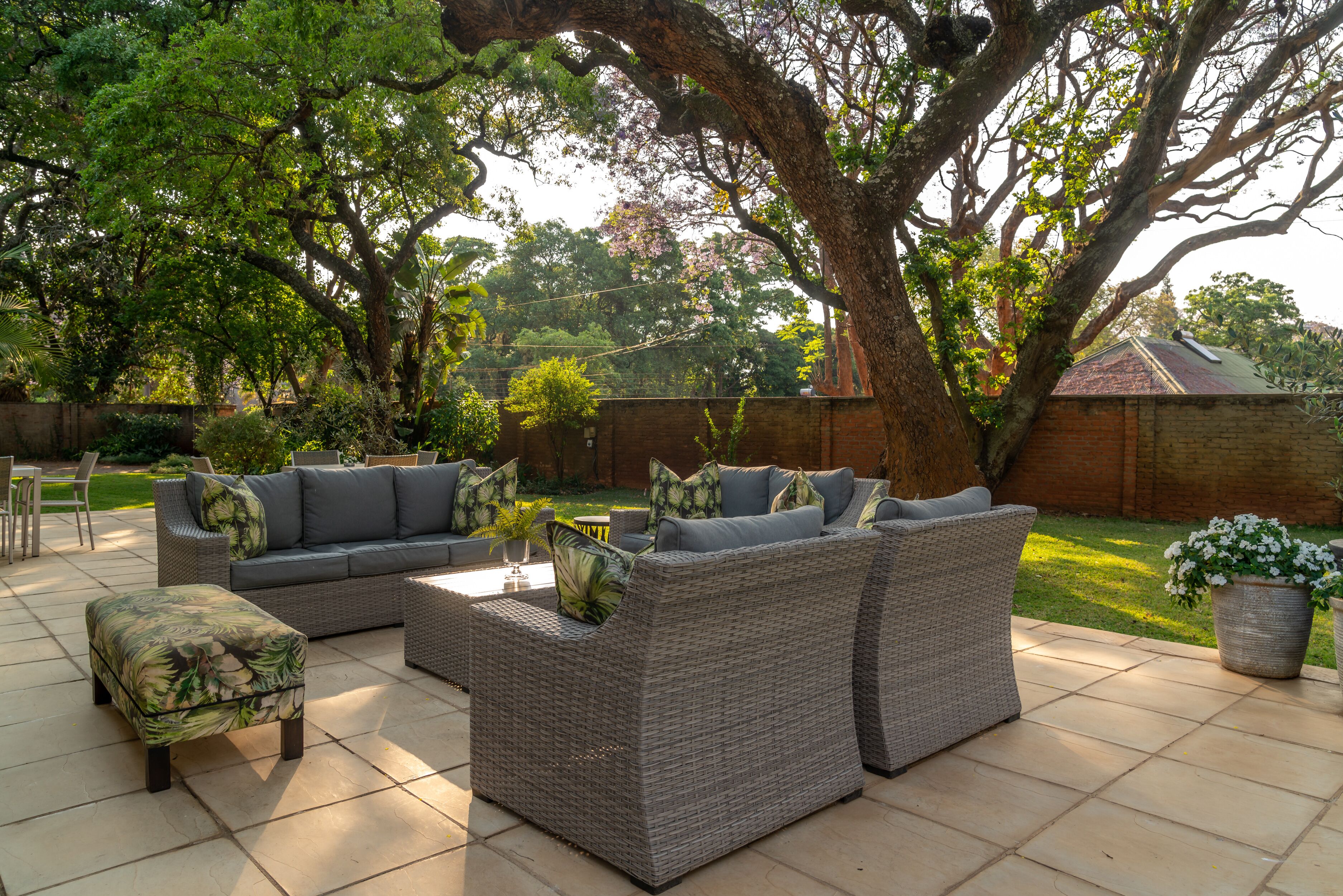 Un patio con mobiliario de mimbre, incluyendo un sofá y reposapiés, rodeado por un gran árbol y un muro de ladrillo.