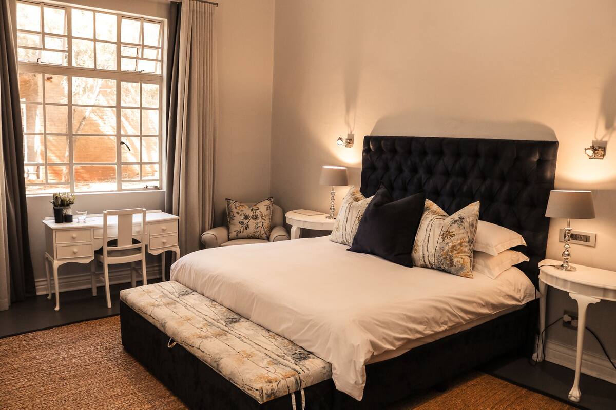 A neatly arranged bedroom with a large bed, bedside tables, a desk, and a window with curtains.