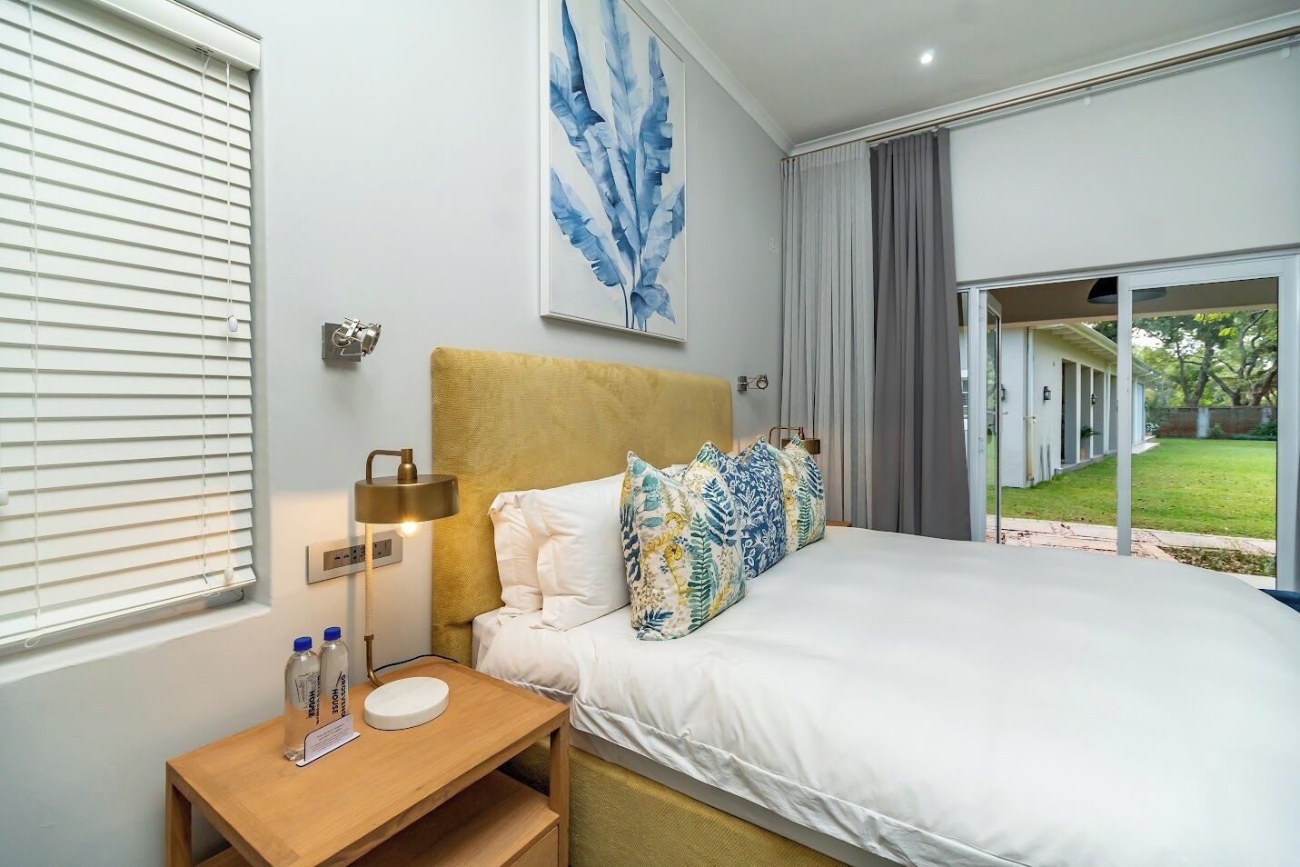 A bedroom with a bed, bedside table, lamp, and a large window with blinds.