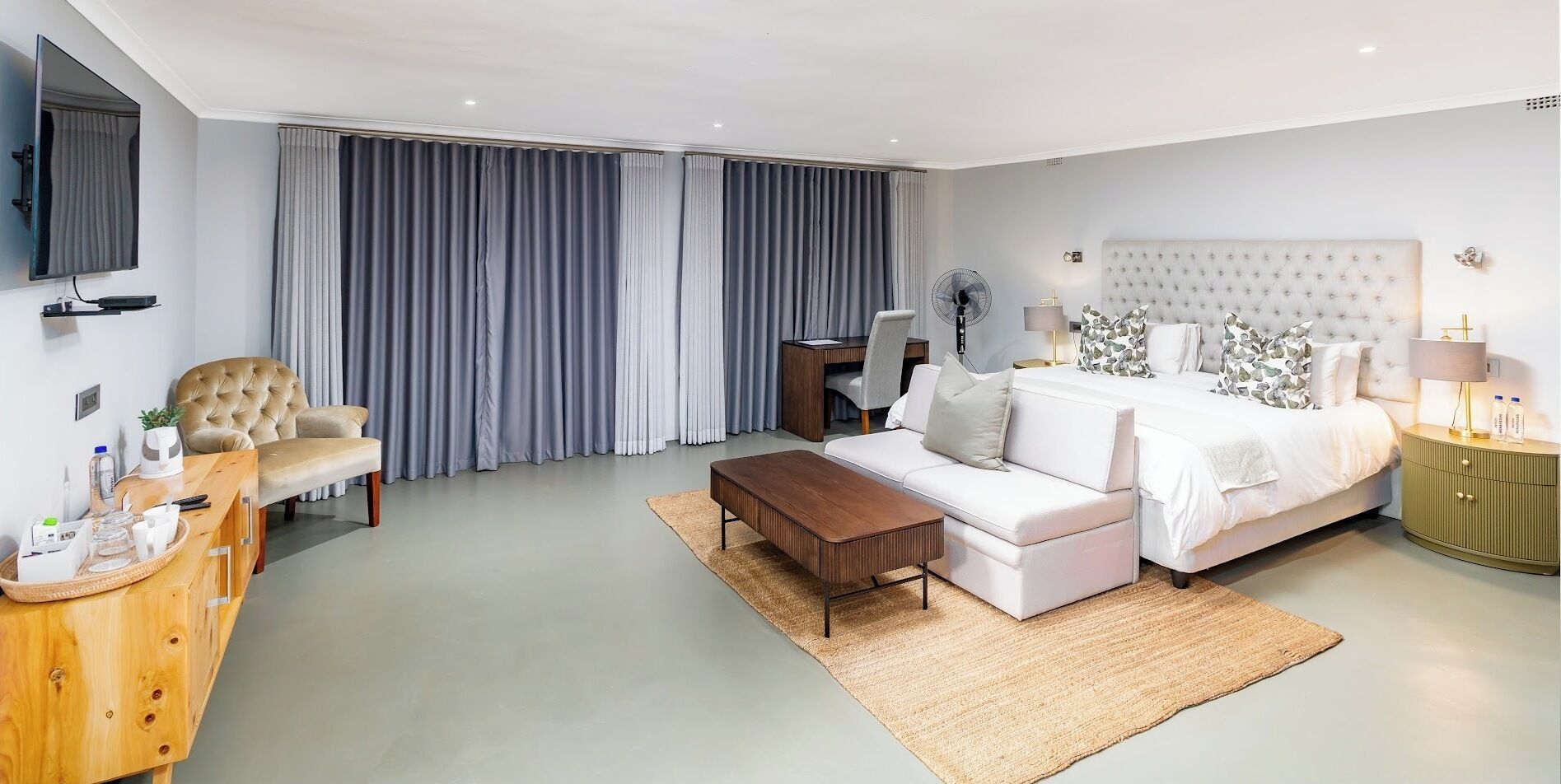 A modern hotel room with a large bed, a sofa, a wooden side table, a desk, and a TV.
