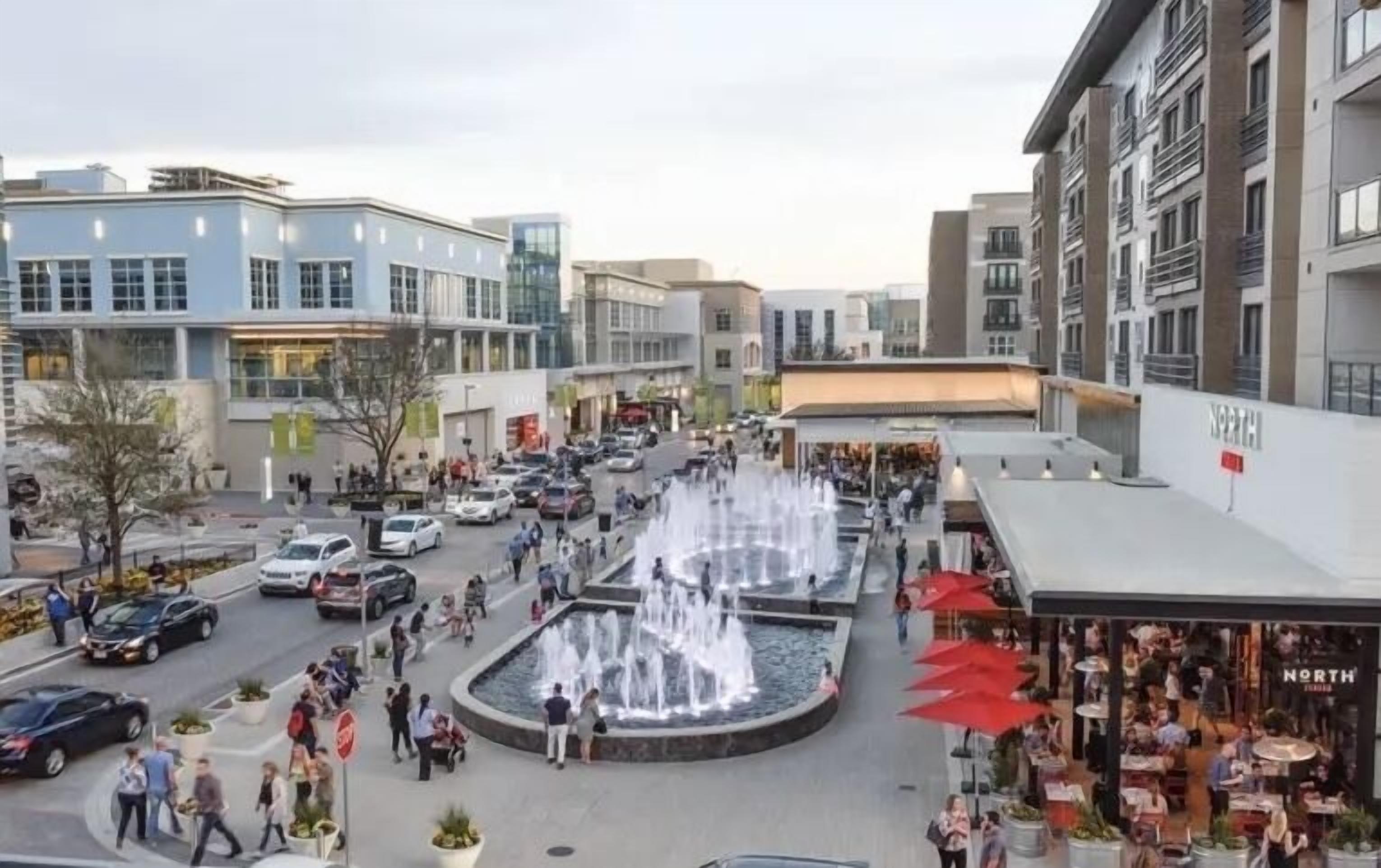 A busy urban square with a central fountain, surrounded by modern buildings and shops.