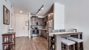 A modern kitchen with stainless steel appliances, a wooden dining table with bar stools, and a hallway leading to a white door. - Plano Tourist Hot Spot! (Plano)
