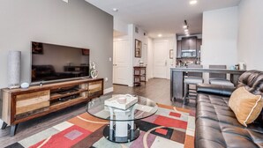 A modern living room with a leather sofa, a glass coffee table, and a flat-screen TV on a wooden console. A kitchen with a bar area is visible in the background. - Plano Tourist Hot Spot! (Plano)