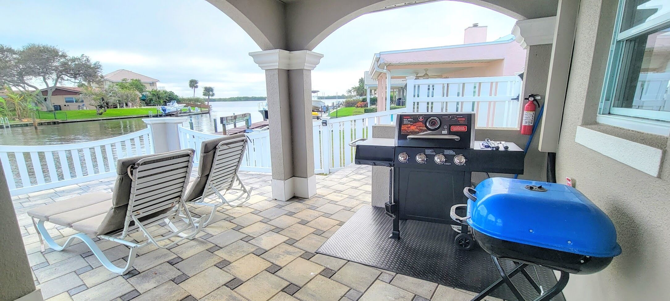 A covered outdoor area with a grill, lounge chairs, and a view of a body of water.