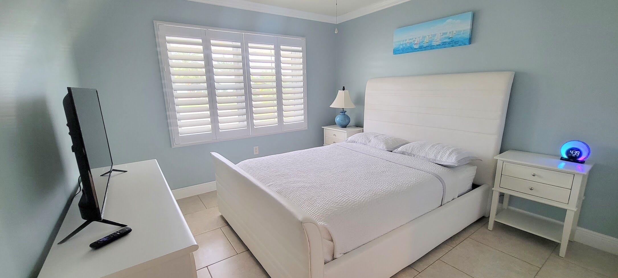 Un dormitorio con cama, escritorio con computadora, mesita de noche con lámpara y un reloj de pared.